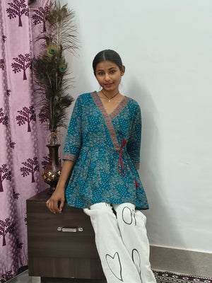 Woman wearing teal blue floral wrap style short cotton kurti indoors standing beside decorative peacock feathers