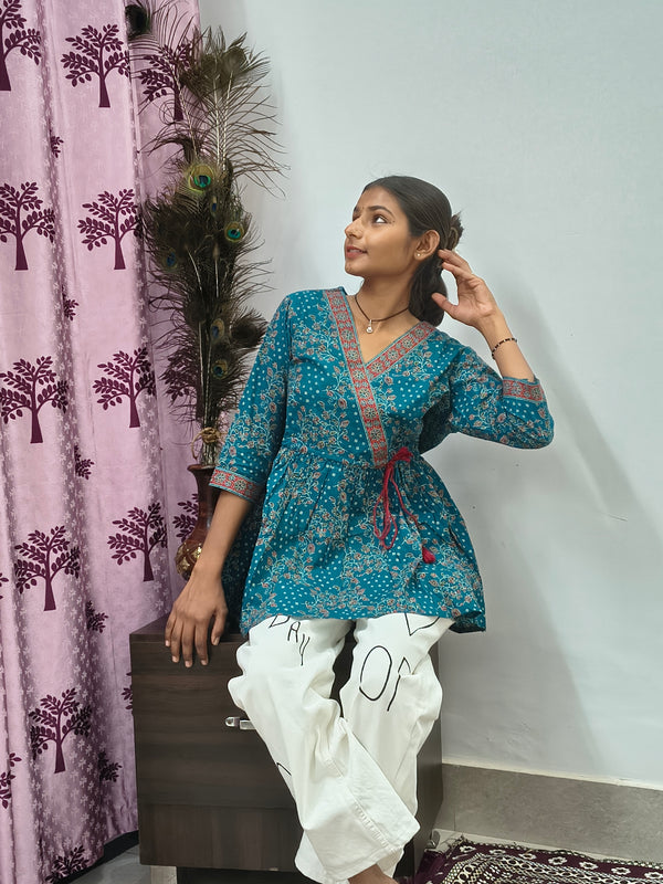 Woman wearing teal blue floral wrap style short cotton kurti indoors standing beside decorative peacock feathers