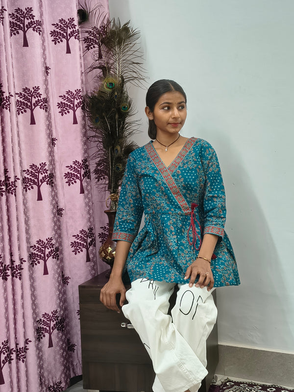 Woman wearing teal blue floral wrap style short cotton kurti indoors standing beside decorative peacock feathers