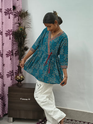 Woman wearing teal blue floral wrap style short cotton kurti indoors standing beside decorative peacock feathers