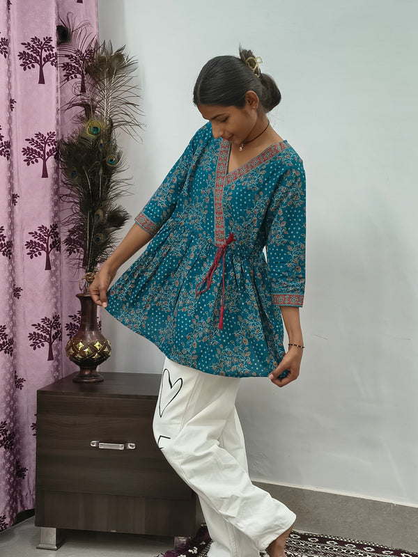Woman wearing teal blue floral wrap style short cotton kurti indoors standing beside decorative peacock feathers