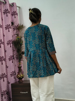 Woman wearing teal blue floral wrap style short cotton kurti indoors standing beside decorative peacock feathers