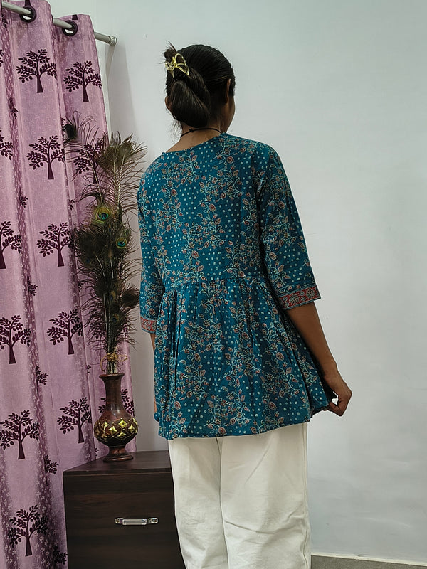 Woman wearing teal blue floral wrap style short cotton kurti indoors standing beside decorative peacock feathers