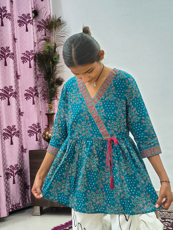 Woman wearing teal blue floral wrap style short cotton kurti indoors standing beside decorative peacock feathers