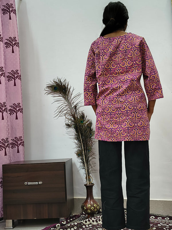Model wearing purple cotton short kurti with paisley print standing indoors next to decorative feathers
