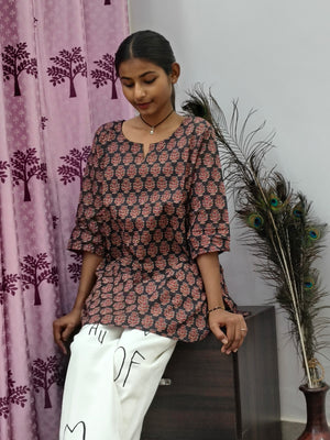 Model wearing black cotton short kurti with rust orange floral block print indoors sitting by cabinet