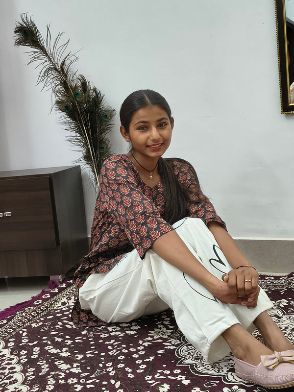 Model wearing black cotton short kurti with rust orange floral block print indoors sitting by cabinet