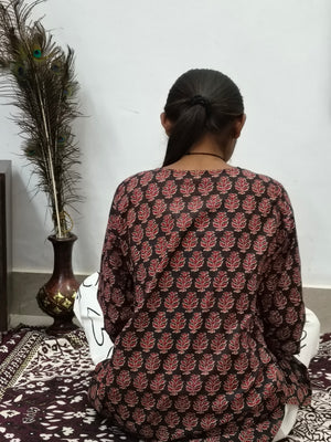 Model wearing black cotton short kurti with rust orange floral block print indoors sitting by cabinet