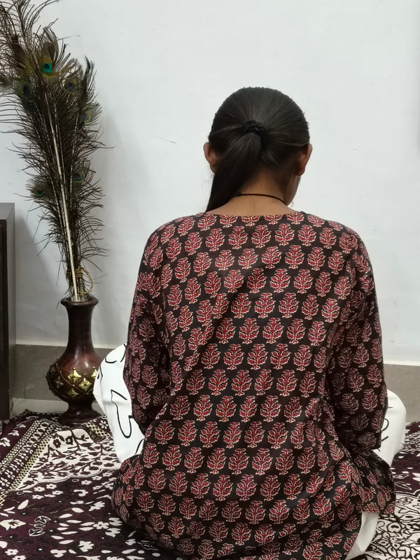 Model wearing black cotton short kurti with rust orange floral block print indoors sitting by cabinet