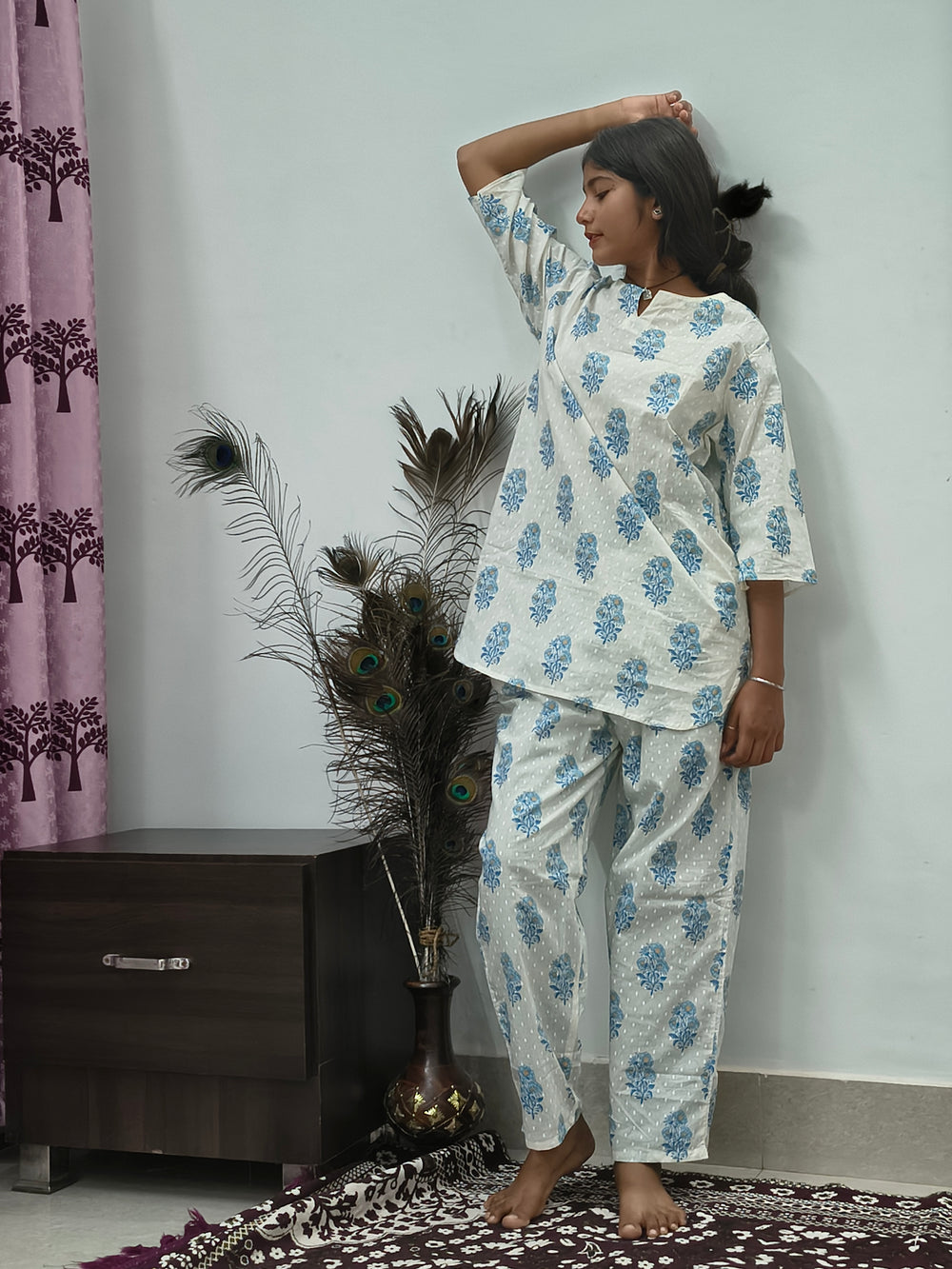 Woman wearing white and blue floral block print cotton kurta pajama set standing indoors with peacock feather decor